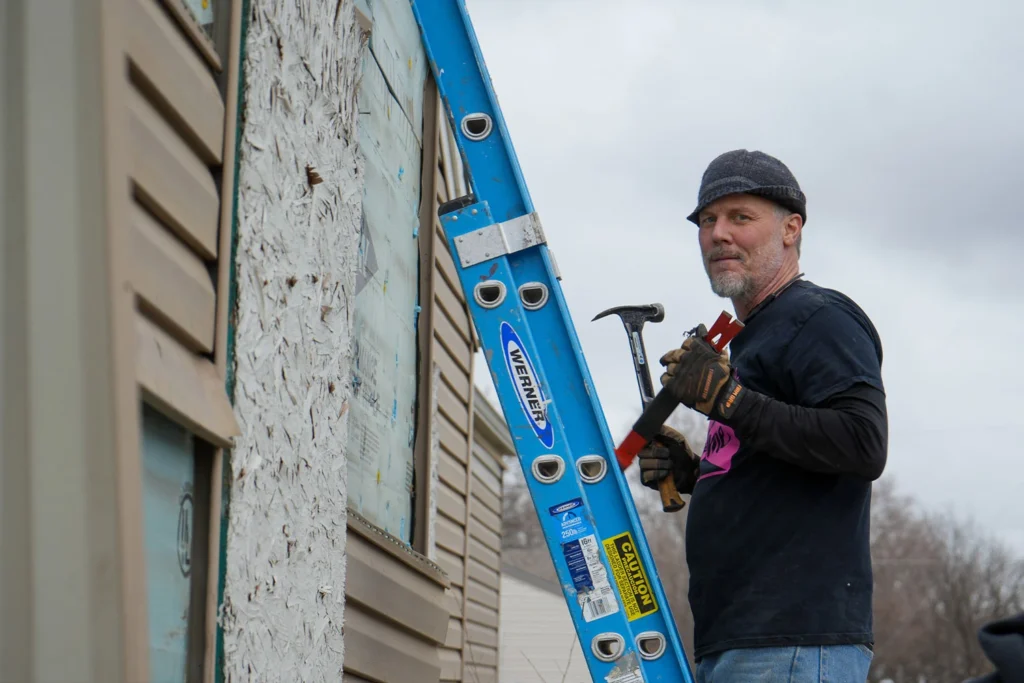 A construction staff taking siding off the wall on a ladder.