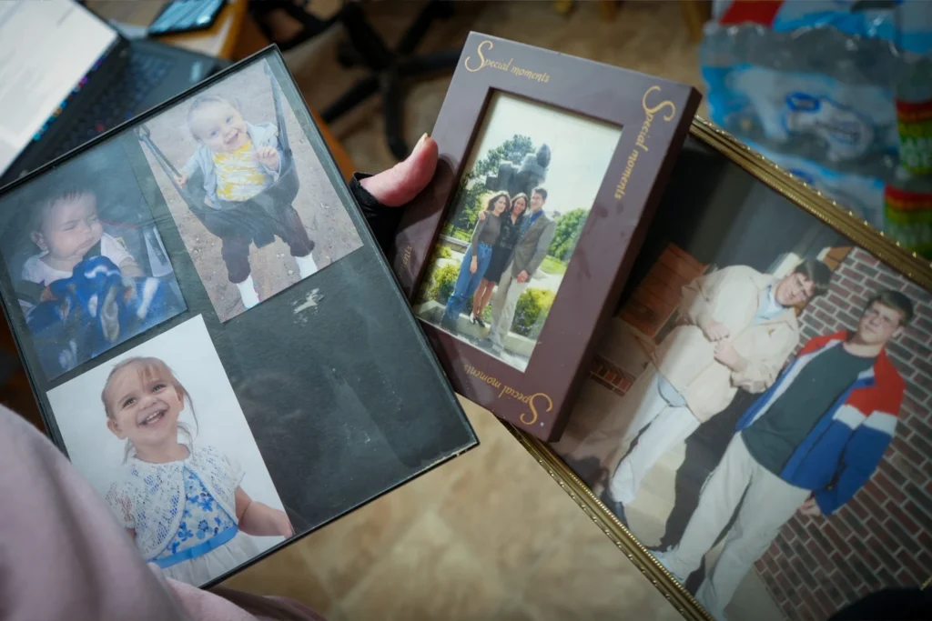 Hands holding a black photo album page displaying three pictures of young children, a brown frame with a photo of three adults, and a gold frame showing two young men.