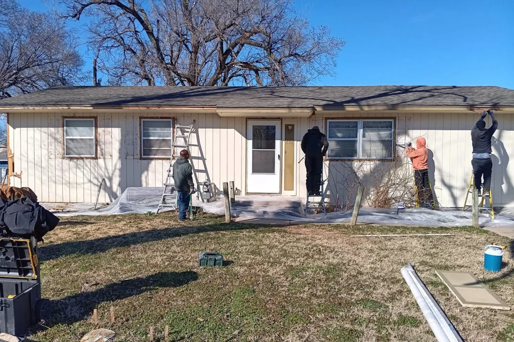 A photo of four workers renovating the exterior of a light beige, single-story house.