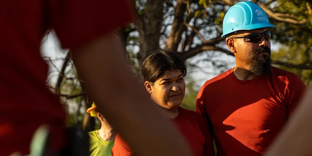 A group of Habitat for Humanity volunteers and homeowners holding hands in a circle during an outdoor build event.