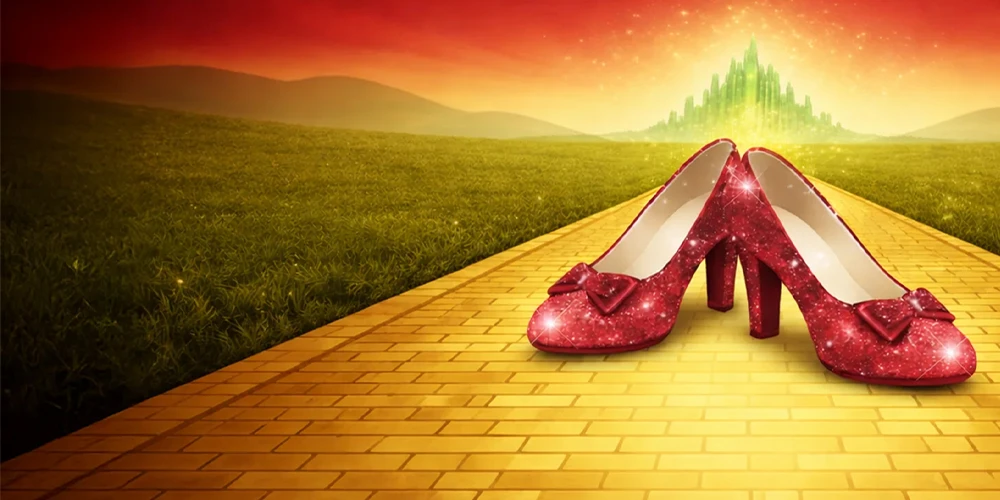 Glittering ruby slippers rest on a yellow brick road with the Emerald City in the back.