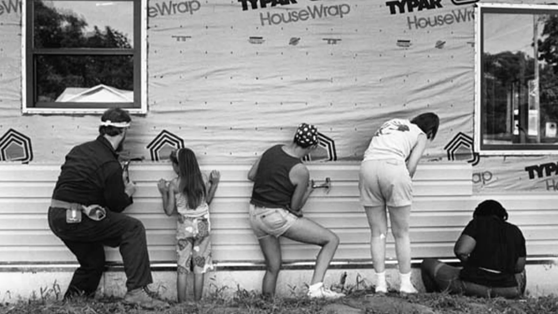 Five volunteers, including a child, kneeling to install exterior siding on a house.