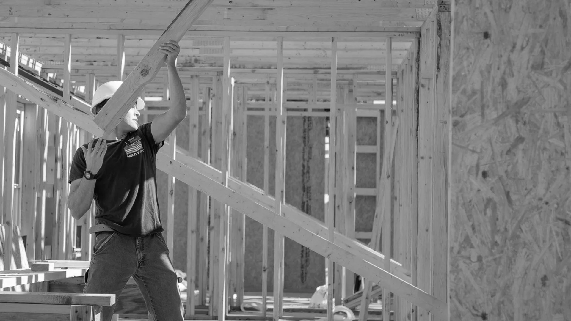 Black and white photo of a volunteer in a hard hat lifting a wooden board inside the frame of a house.