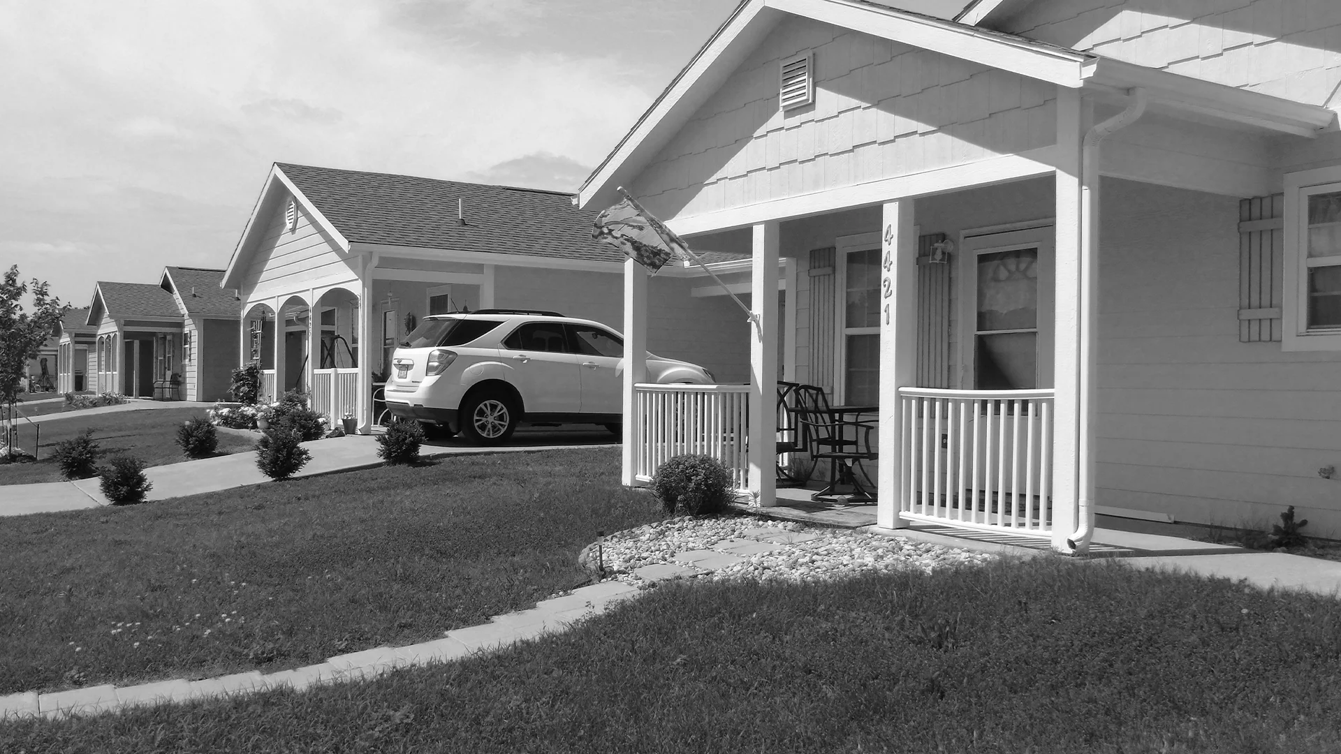 Black and white photo of a row of completed single-story Habitat homes with porches and a white SUV parked in a driveway.