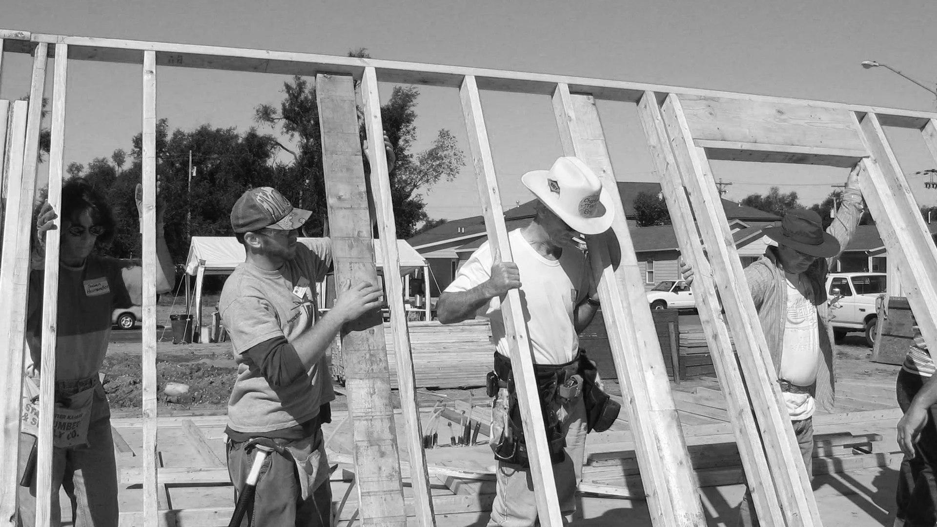 Black and white photo of a group of volunteers, including one in a cowboy hat, lifting a wooden wall frame into place at a construction site.