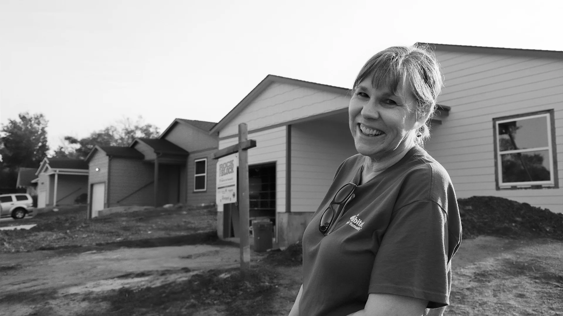 Black and white photo of a smiling woman in a Habitat t-shirt standing in front of several homes under construction.