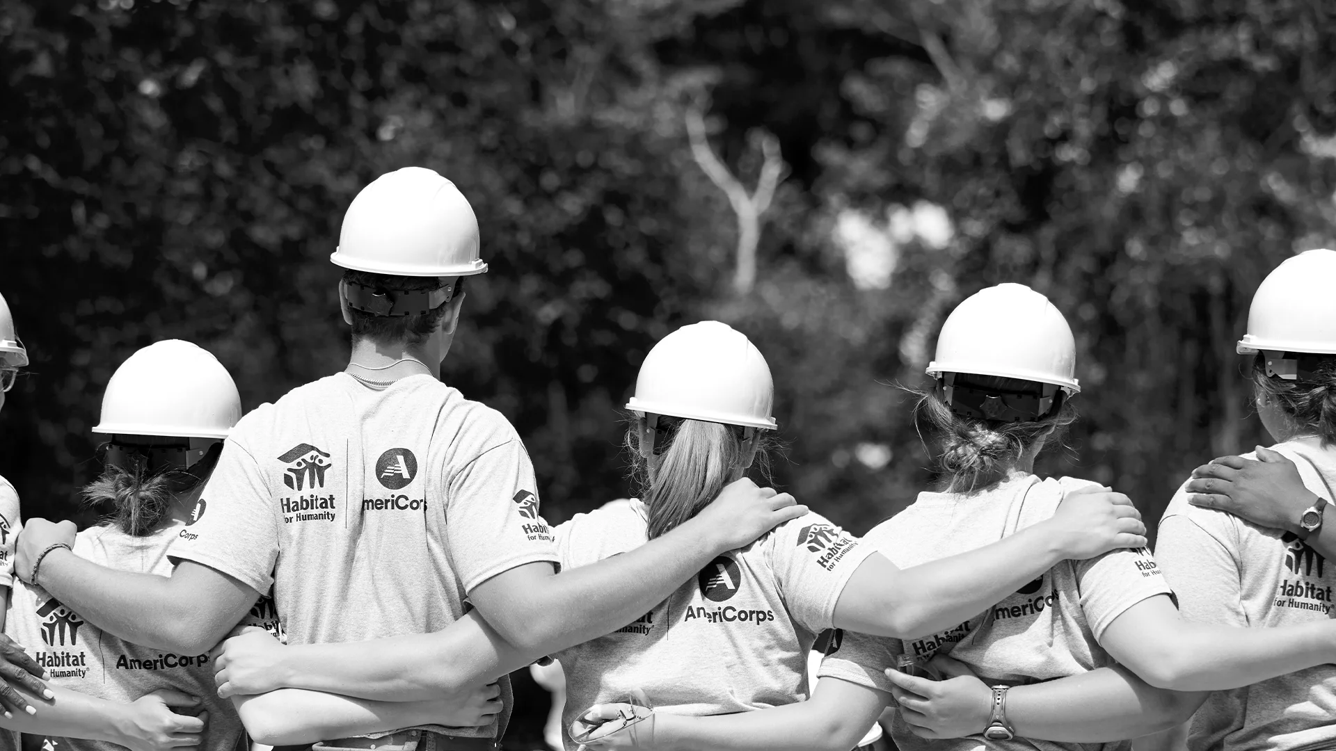 Back view of six AmeriCorps members wearing hard hats and matching Habitat t-shirts, standing with their arms linked around each other.