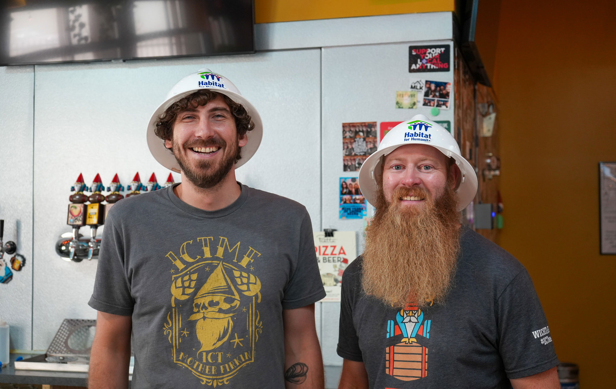 Two men smiling together while wearing Habitat for Humanity hard hats.