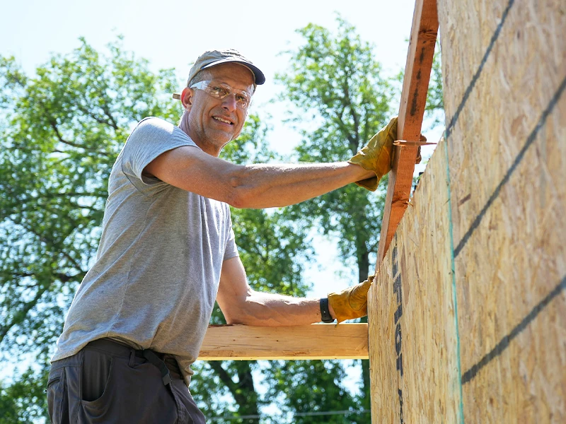 A volunteer smiling while holding onto the siding of a home.