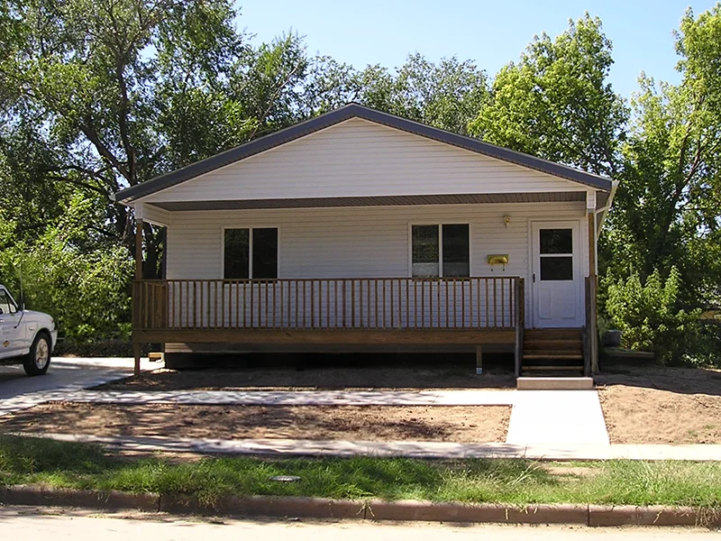 A small, single-story white house with a wooden front porch and steps, surrounded by green trees.