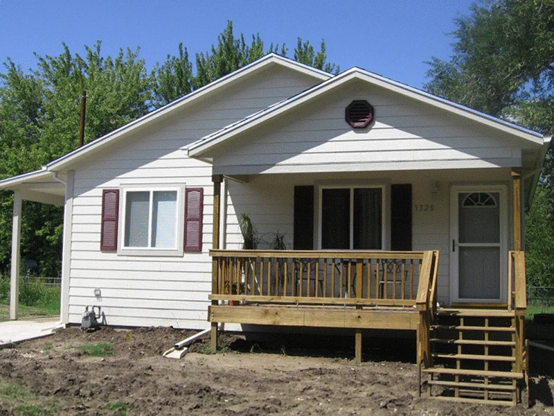 A small, single-story white house with a wooden front porch and steps, surrounded by green trees.