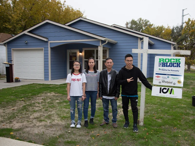 A small, single-story, blue house with a garage and a family standing in on the lawn.