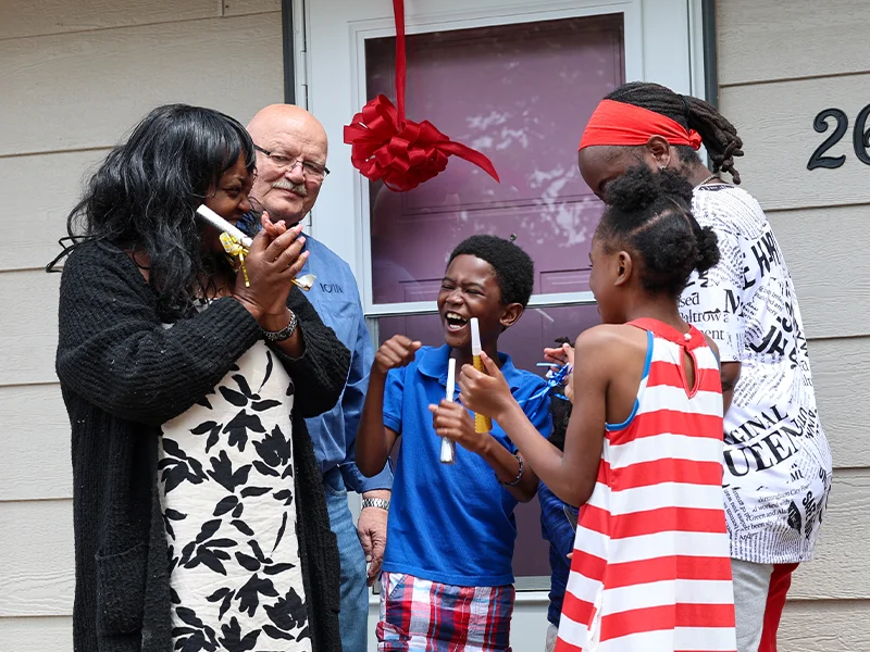 A multi-generational family celebrating outside a home with a red ribbon on the door.