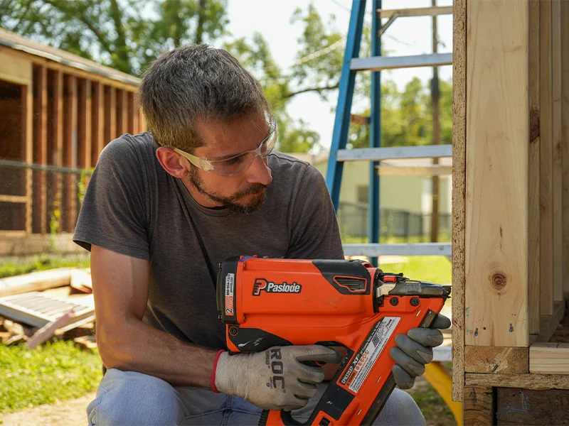 Construction volunteer wearing safety glasses using a nail gun to secure a wooden frame.